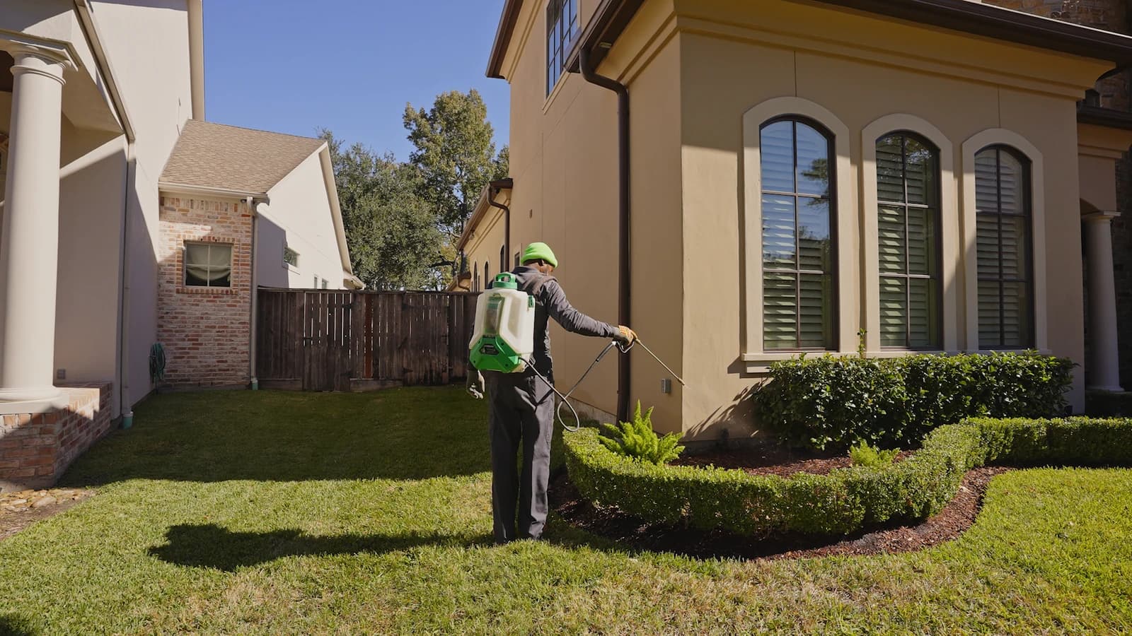 Pest-control technician spraying a liquid treatment along a home foundation and the soil line